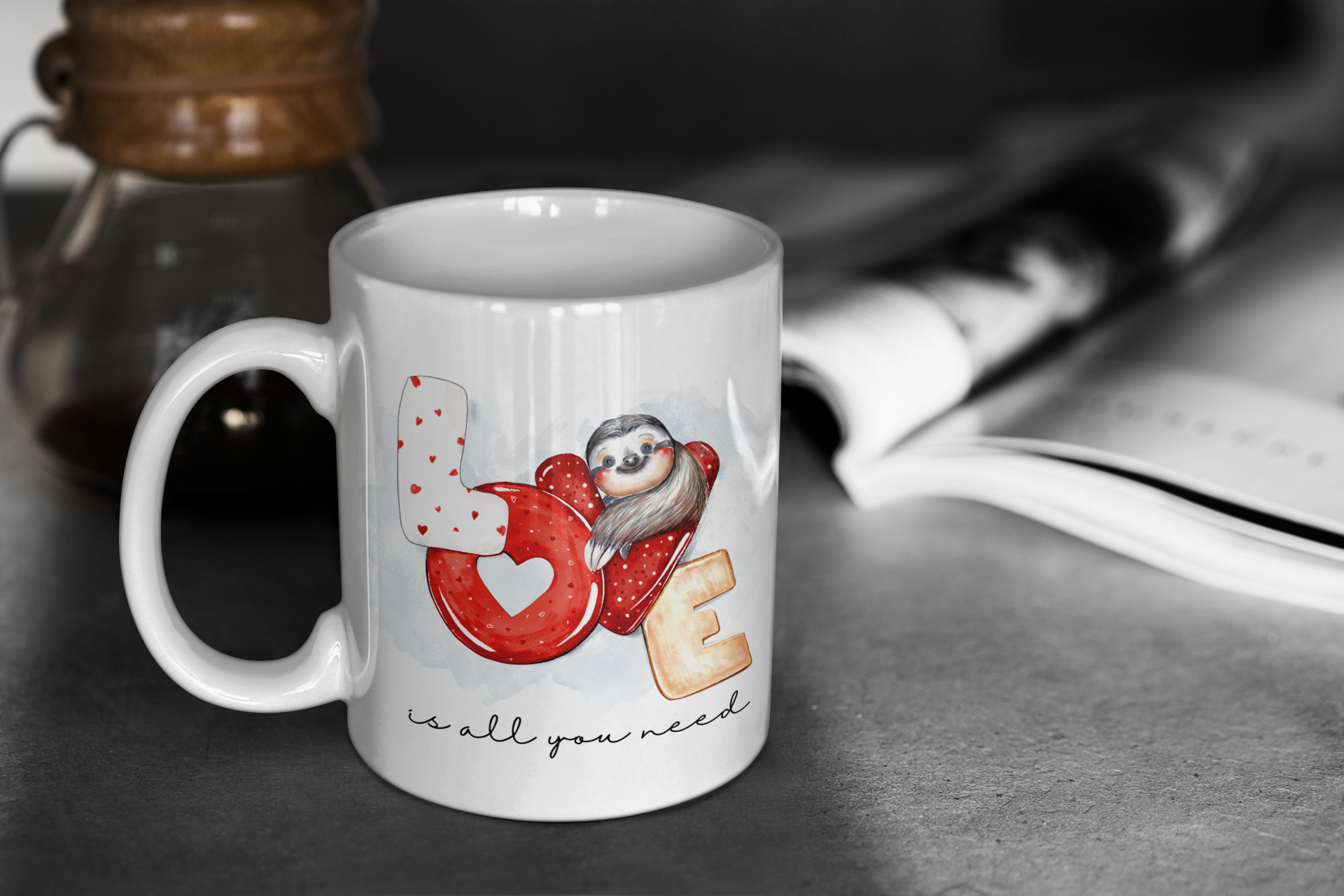 White mug with a cartoon design of a sloth on a heart, with 'LOVE is all you need' text, on a blurred background.