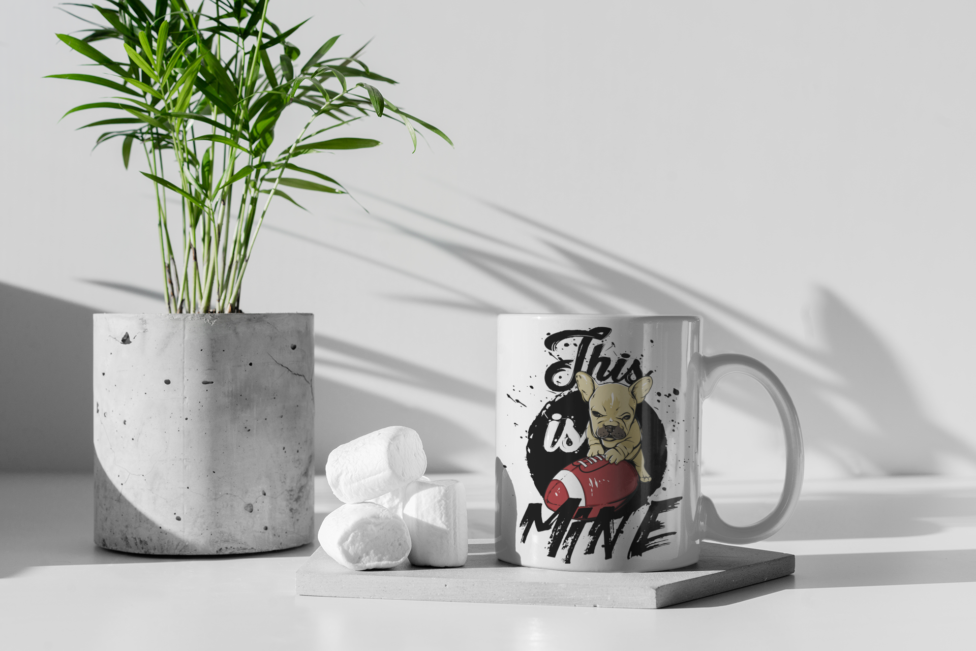 A white ceramic coffee mug with a graphic print of a French bulldog and the text 'This is MINE' sits on a gray tray next to white marshmallows, with a potted plant in the background.