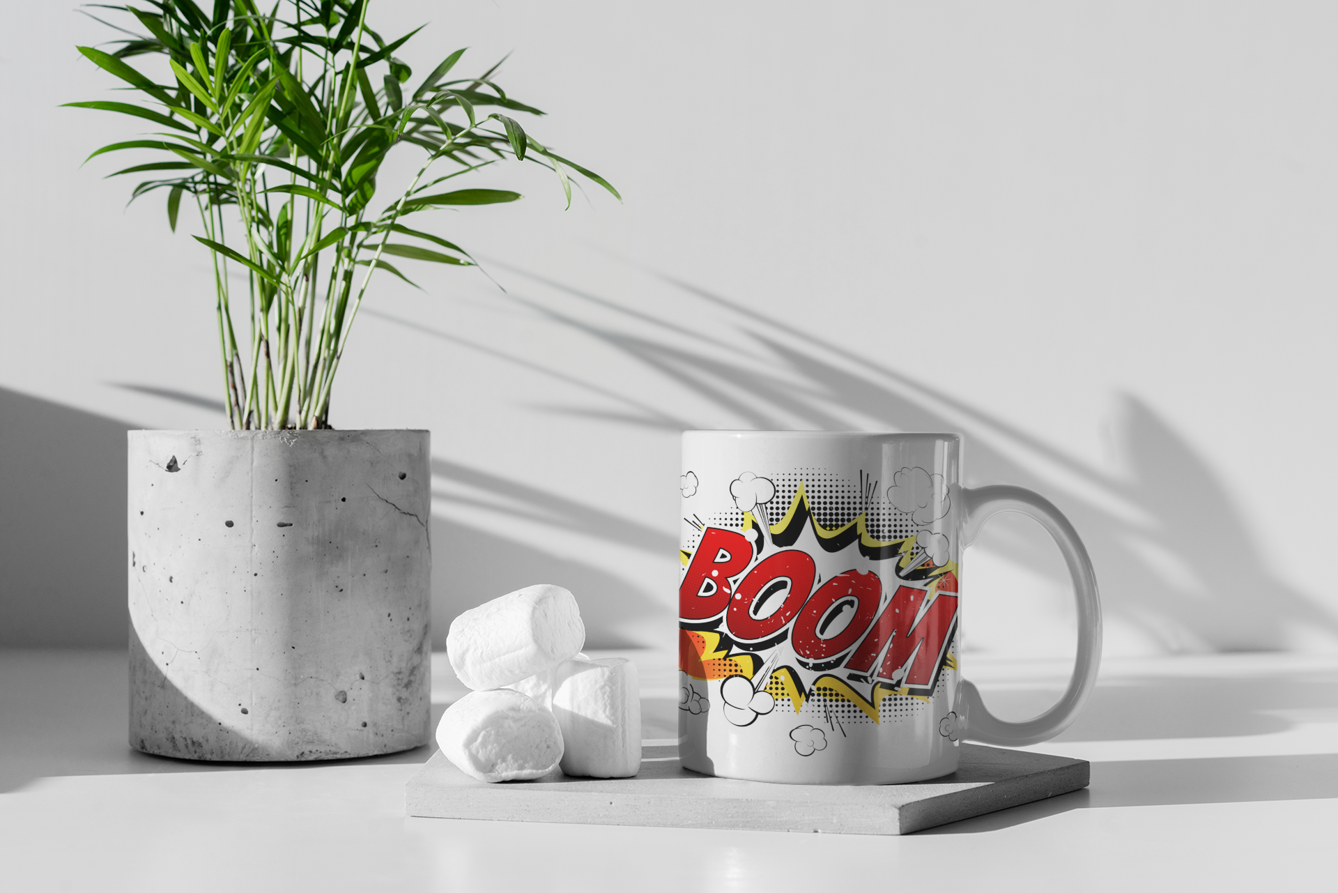 A white ceramic mug with a comic-style 'BOOM' graphic sits on a small tray beside a potted plant and marshmallows in a minimalist studio setting.