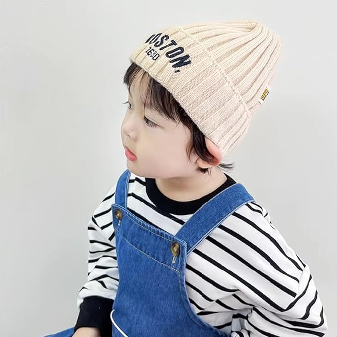 Child wearing a beige knit hat with text, striped shirt, and blue overalls on a light gray background