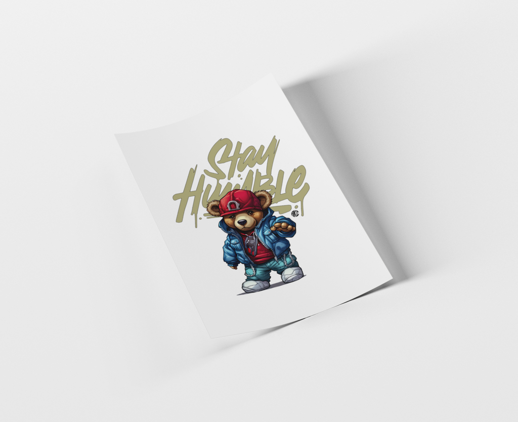 A printed card or poster shown at an angle on a light gray background. The card features a cartoon teddy bear wearing a red cap and blue jacket beneath the words 'Stay Humble' in a gold graffiti-style font.