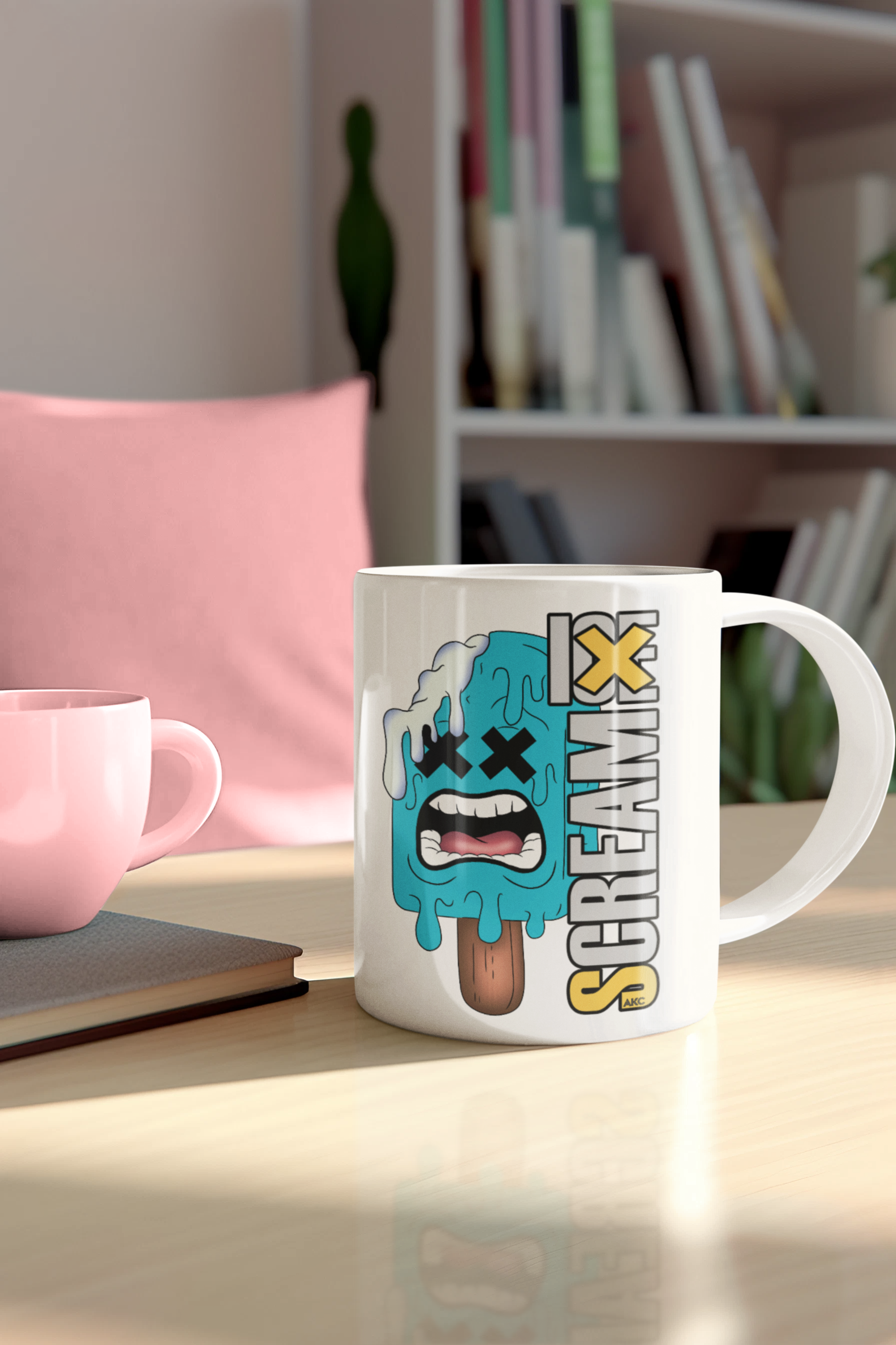 A white ceramic coffee mug with a cartoon graphic of a crying popsicle and the word 'SCREAM' printed on it, sitting on a wooden table in a cozy room setting with a pink mug and bookshelf in the background.