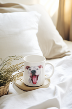 A white ceramic mug with a printed illustration of a white dog with its tongue out, resting on a small beige coaster on a bed with white sheets and pillows.