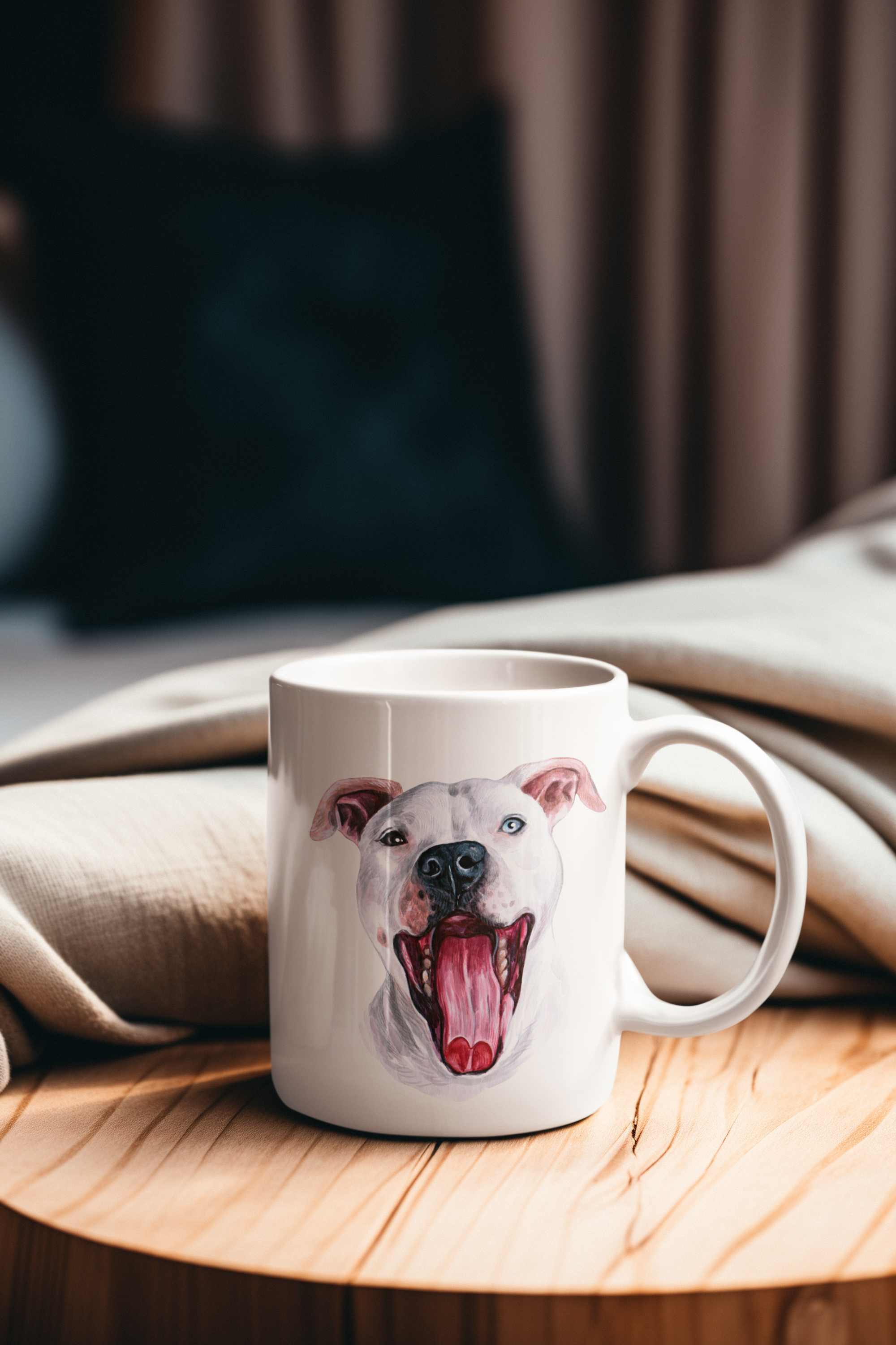 A white ceramic mug featuring a painted portrait of a white dog with its mouth open, shown on a round wooden table with a beige blanket in the background.