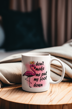 A white ceramic mug with a pink flamingo illustration and the text 'don't make me put my foot down' in black script, set on a wooden table with a beige blanket in the background.