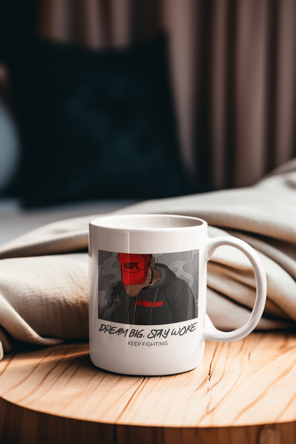 A white ceramic mug with a custom graphic print of a person in a black hoodie and red cap, sitting on a round wooden table with a beige blanket in the background.