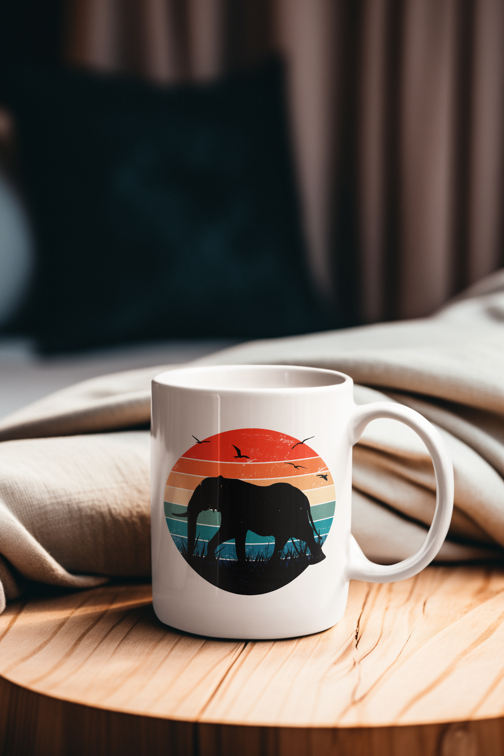 A white ceramic mug with a colorful retro sunset graphic featuring a black elephant silhouette, placed on a round wooden table with a beige blanket and a blurred indoor background.