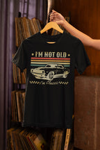 Black t-shirt with car graphic and text 'I'm not old, I'm classic' on a hanger against a bookshelf background.