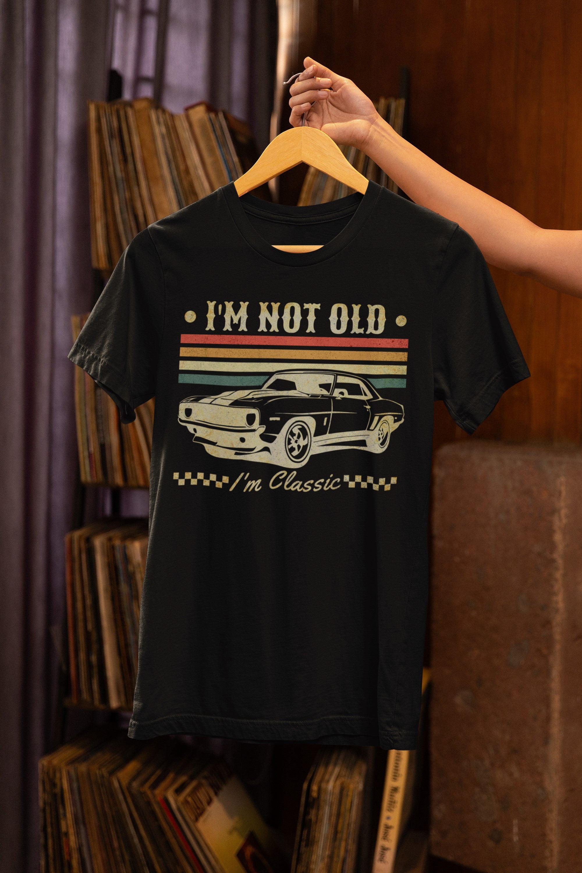 Black t-shirt with car graphic and text 'I'm not old, I'm classic' on a hanger against a bookshelf background.