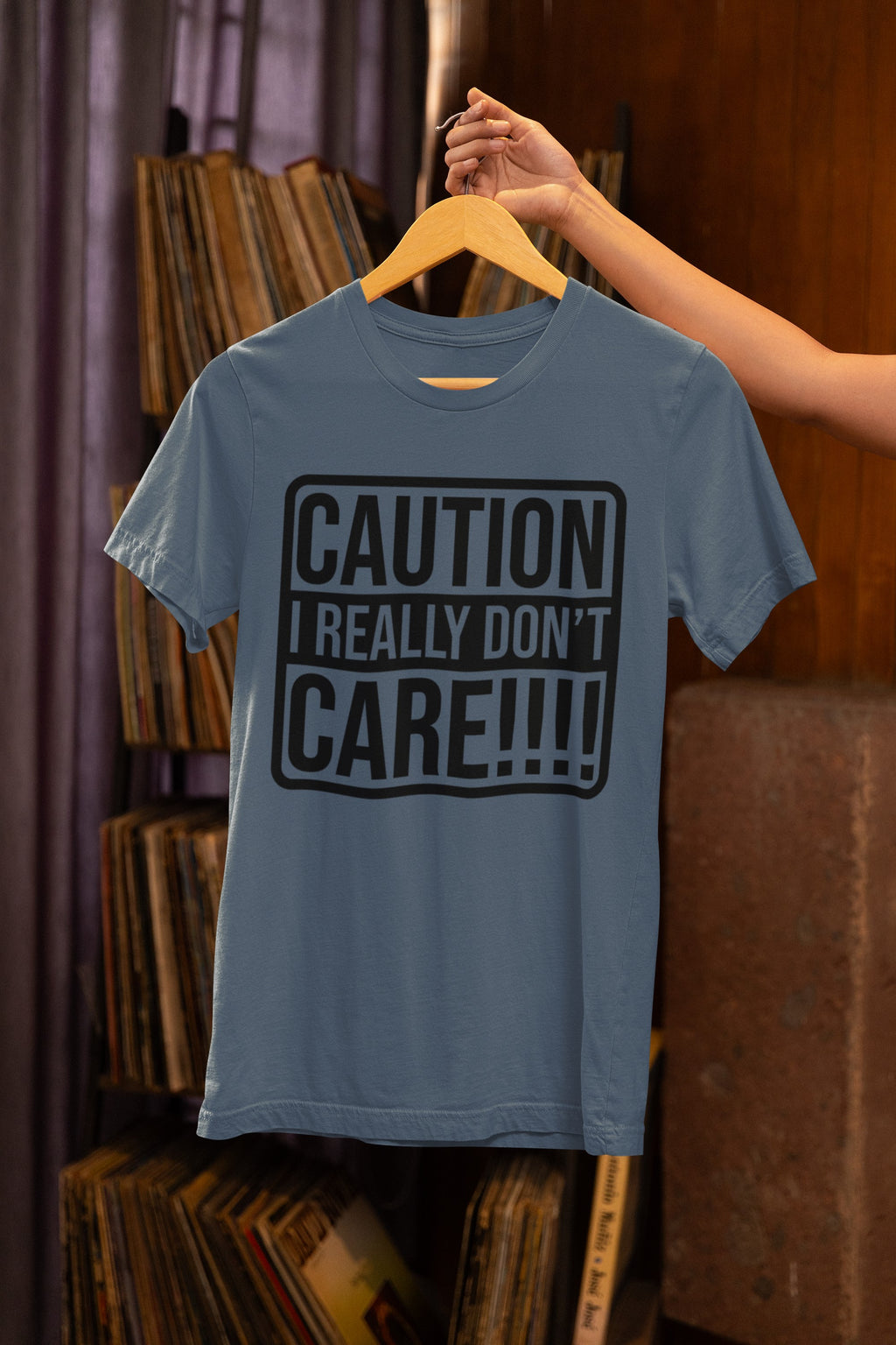 Blue t-shirt with 'CAUTION I REALLY DON'T CARE!!!' text on a hanger against a bookshelf background.