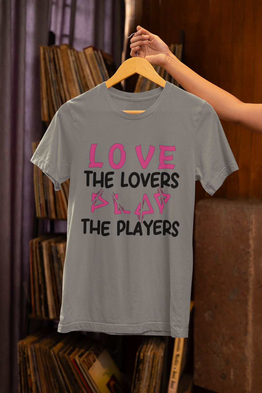 Gray t-shirt with pink and black text on a hanger against a bookshelf background
