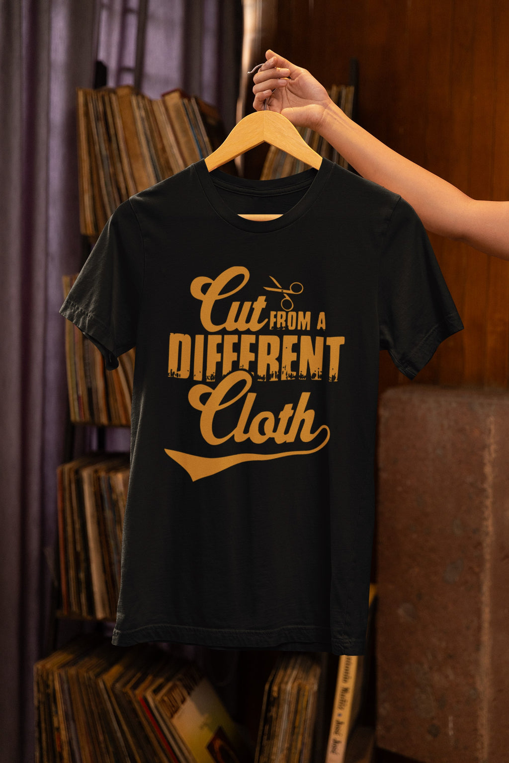 Black t-shirt with yellow text 'Cut From A Different Cloth' on a hanger against a bookshelf background.