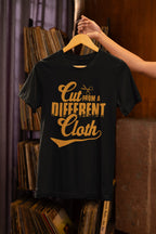 Black t-shirt with yellow text 'Cut From A Different Cloth' on a hanger against a bookshelf background.