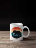 A white ceramic mug with a colorful retro sunset graphic featuring an elephant silhouette, placed on a wooden surface against a black background.