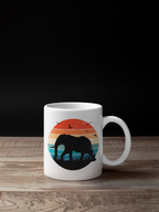 A white ceramic mug with a colorful retro sunset graphic featuring an elephant silhouette, placed on a wooden surface against a black background.