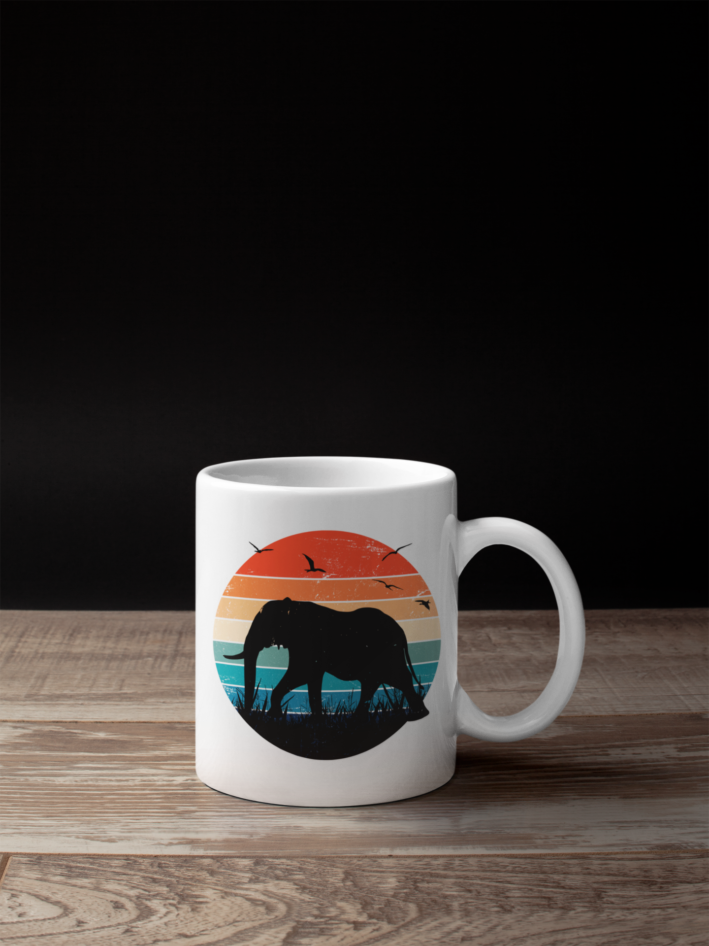 A white ceramic mug with a colorful retro sunset graphic featuring an elephant silhouette, placed on a wooden surface against a black background.
