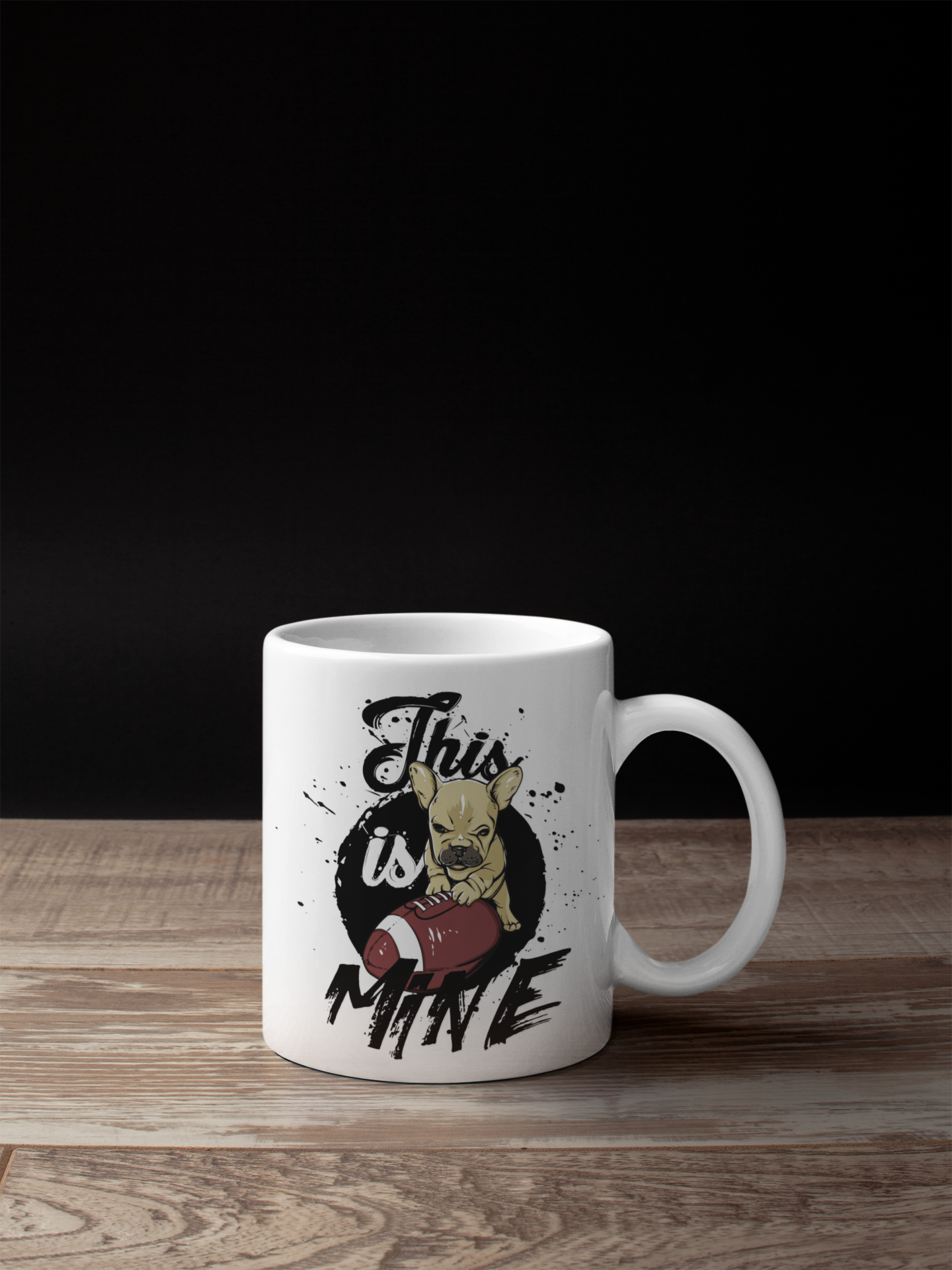 A white ceramic coffee mug with a custom graphic print of a French Bulldog holding a football and the text 'This is Mine', displayed on a wooden surface against a solid black background.