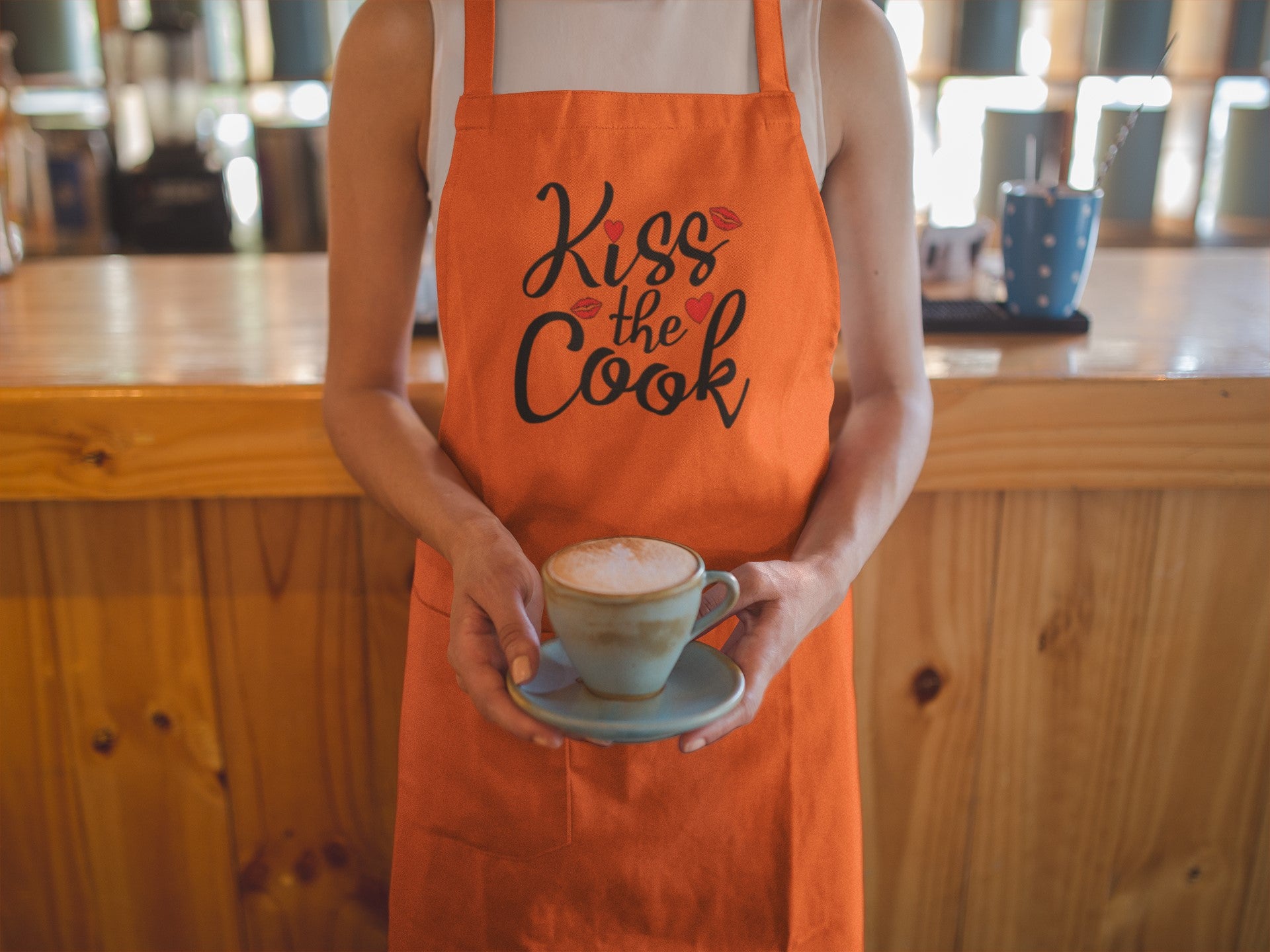 Person wearing an orange apron with 'Kiss the Cook' text, holding a cup of coffee.