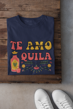 A navy blue t-shirt with a colorful retro graphic reading 'TE AMO QUILA' is laid flat on a rustic wooden surface. The graphic features a pink tequila bottle, a pink drink glass, yellow flowers, and green leaves.