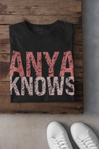 Black t-shirt with 'ANYA KNOWS' text on a wooden surface with white sneakers.