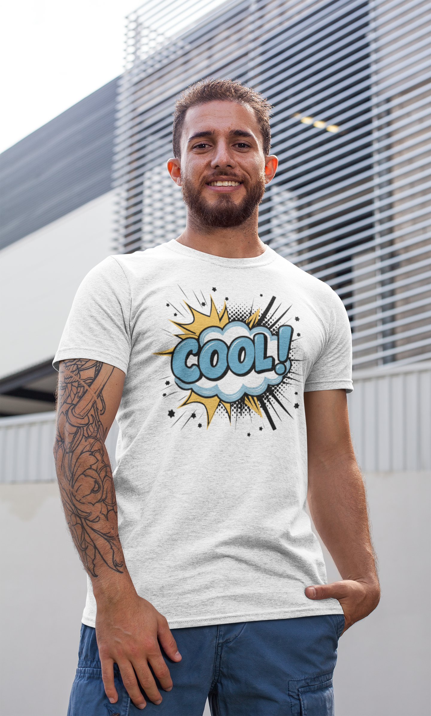 A man wearing a white graphic t-shirt with a blue and yellow comic-style 'COOL!' design, standing in front of a modern building with horizontal slats.