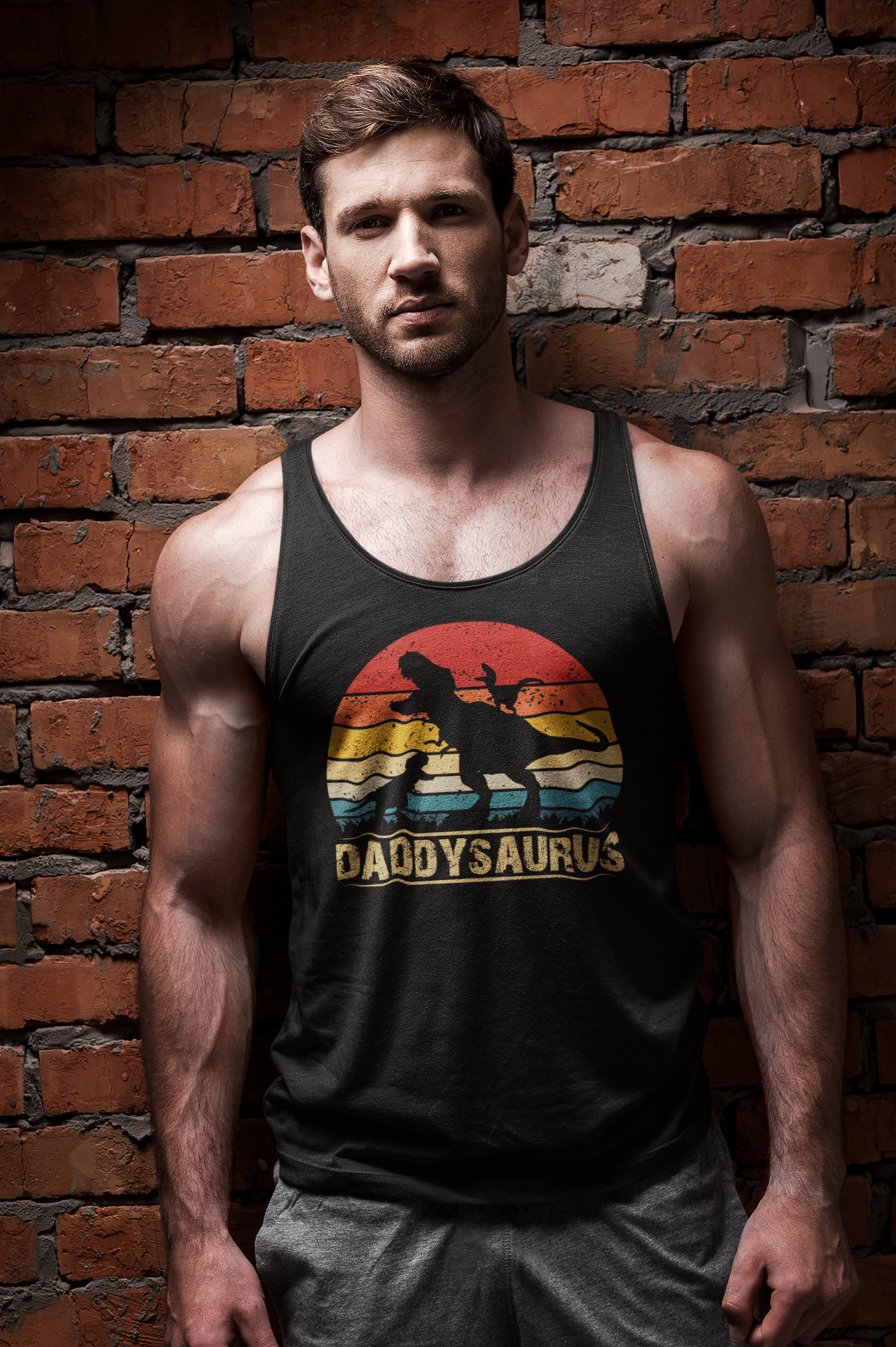A muscular man wearing a black tank top with a retro sunset graphic and the text 'DADDYSAUROS' stands against a red brick wall background.