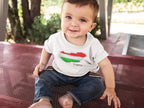 Baby wearing a shirt with a flag design sitting on a metal bench outdoors.