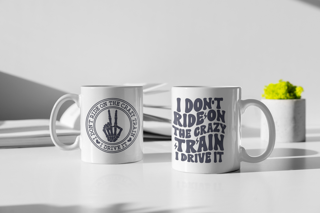 Two white ceramic mugs on a white tabletop in a styled studio setting. The left mug shows a circular black graphic of a hand making the peace sign with the text 'DON'T RIDE ON THE CRAZY TRAIN I DRIVE IT'.