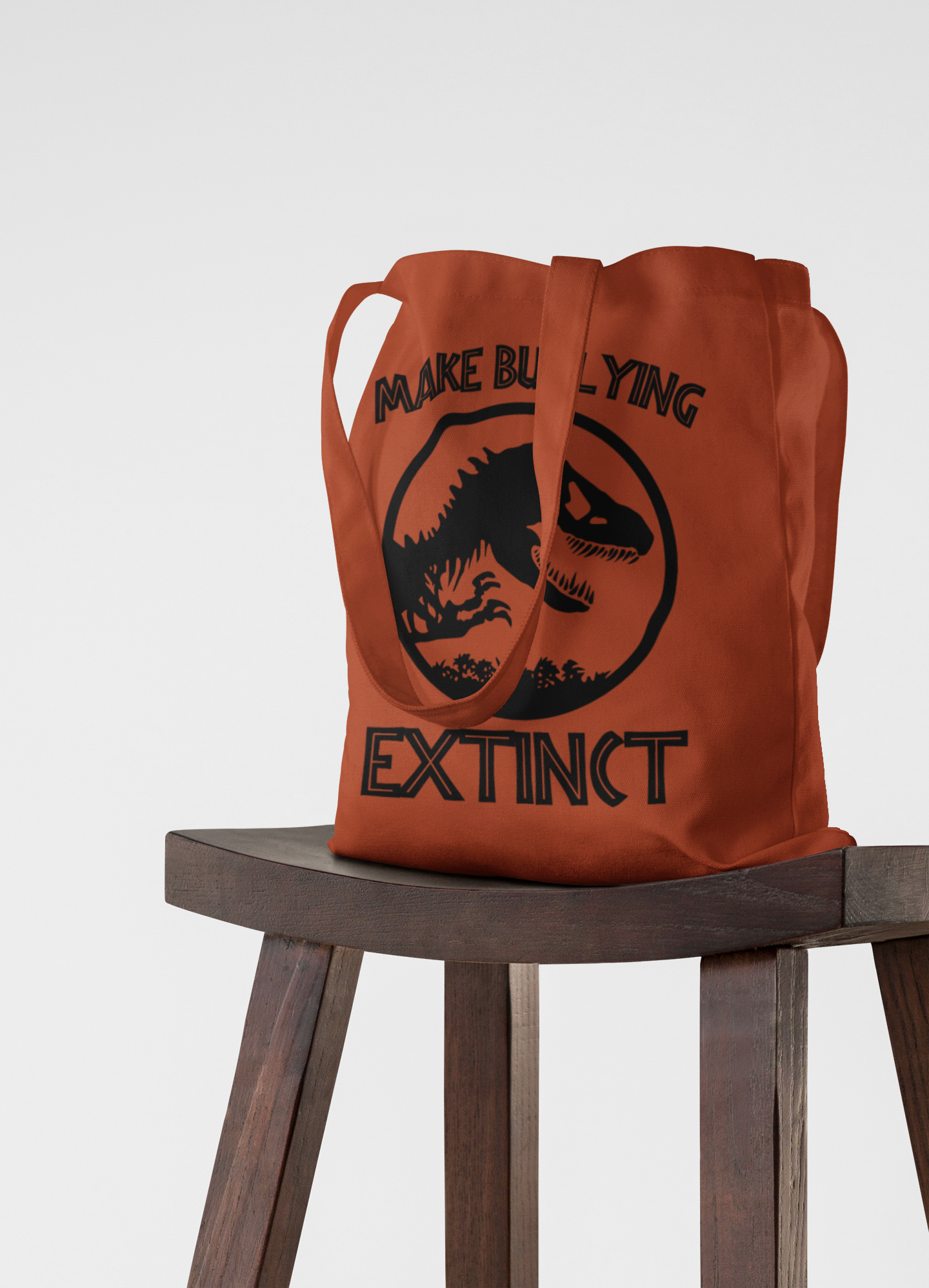 Brown tote bag with dinosaur graphic and text on a wooden stool against a white background