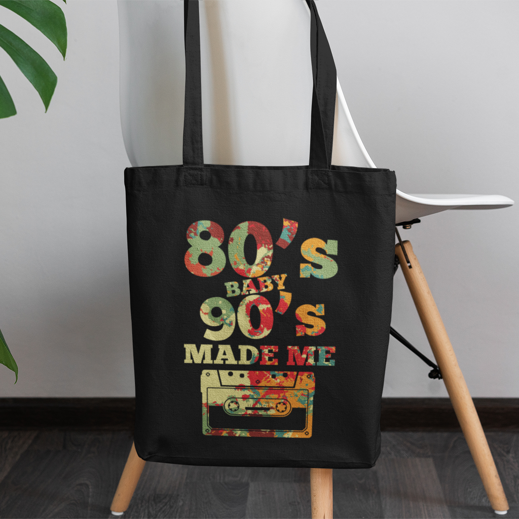 Black tote bag with '80s Baby 90s Made Me' design on a wooden stool with a plant in the background