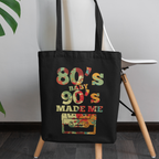 Black tote bag with '80s Baby 90s Made Me' design on a wooden stool with a plant in the background