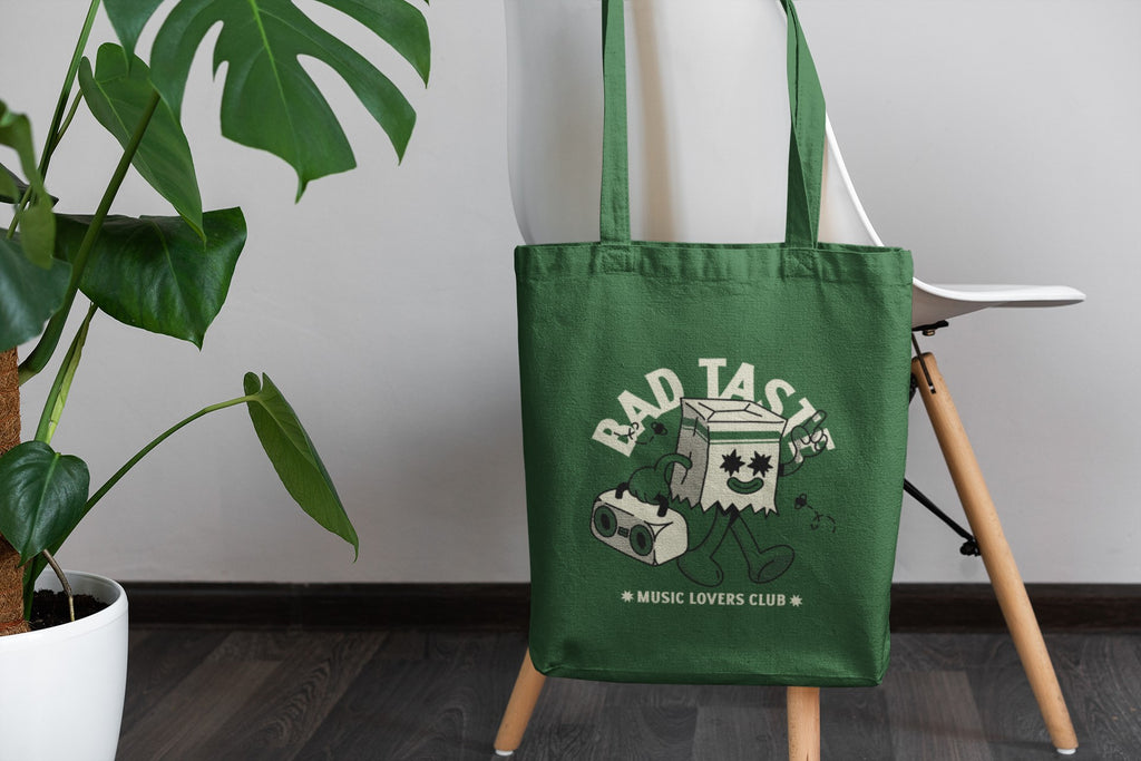 Green tote bag with 'Bad Taste Music Lovers Club' design on a stand next to a plant.