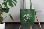 Green tote bag with 'Bad Taste Music Lovers Club' design on a stand next to a plant.
