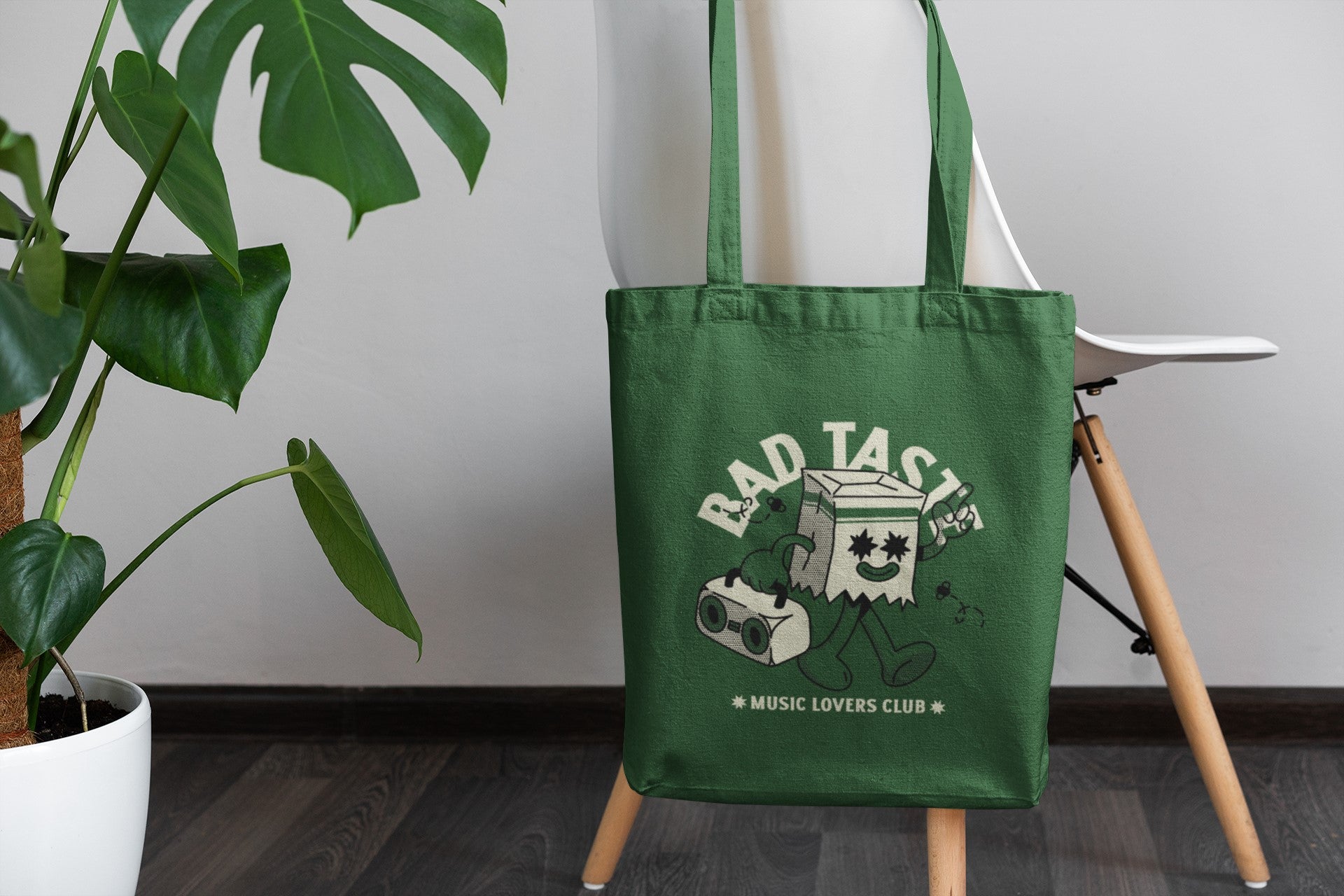 Green tote bag with 'Bad Taste Music Lovers Club' design on a stand next to a plant.