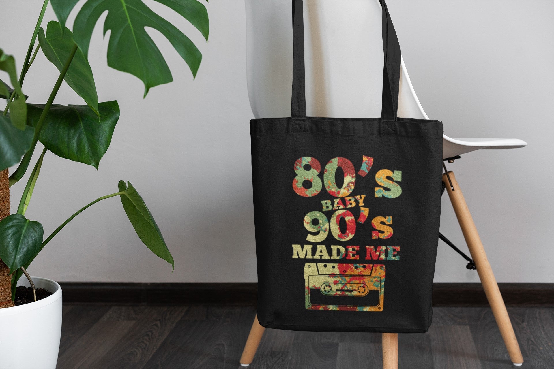 Black tote bag with '80s Baby 90s Made Me' design on a wooden stand with a plant in the background.