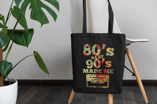Black tote bag with '80s Baby 90s Made Me' design on a wooden stand with a plant in the background.
