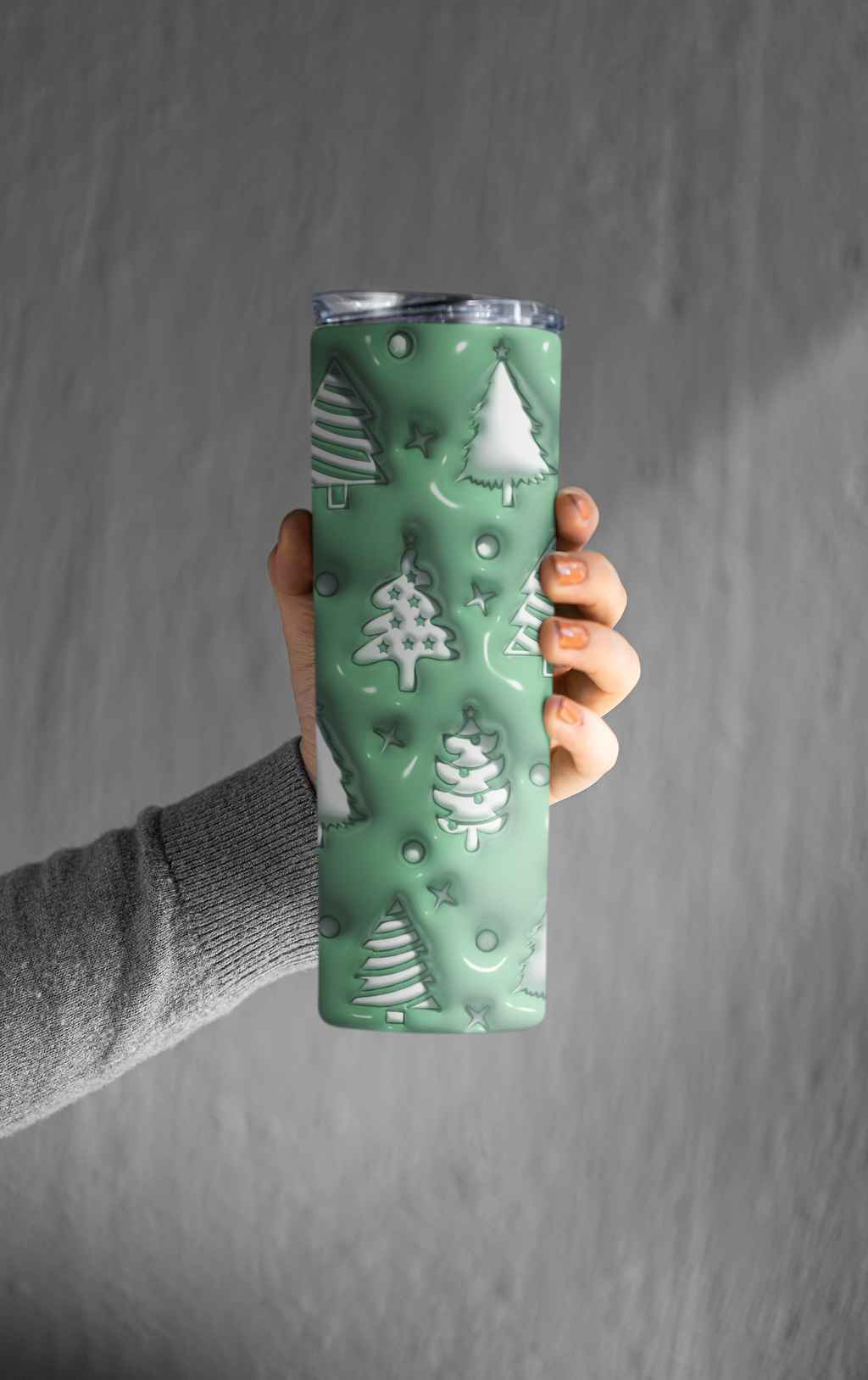 A hand holding a tall insulated tumbler with a mint green holiday design featuring embossed Christmas trees and snowflakes, set against a textured gray background.