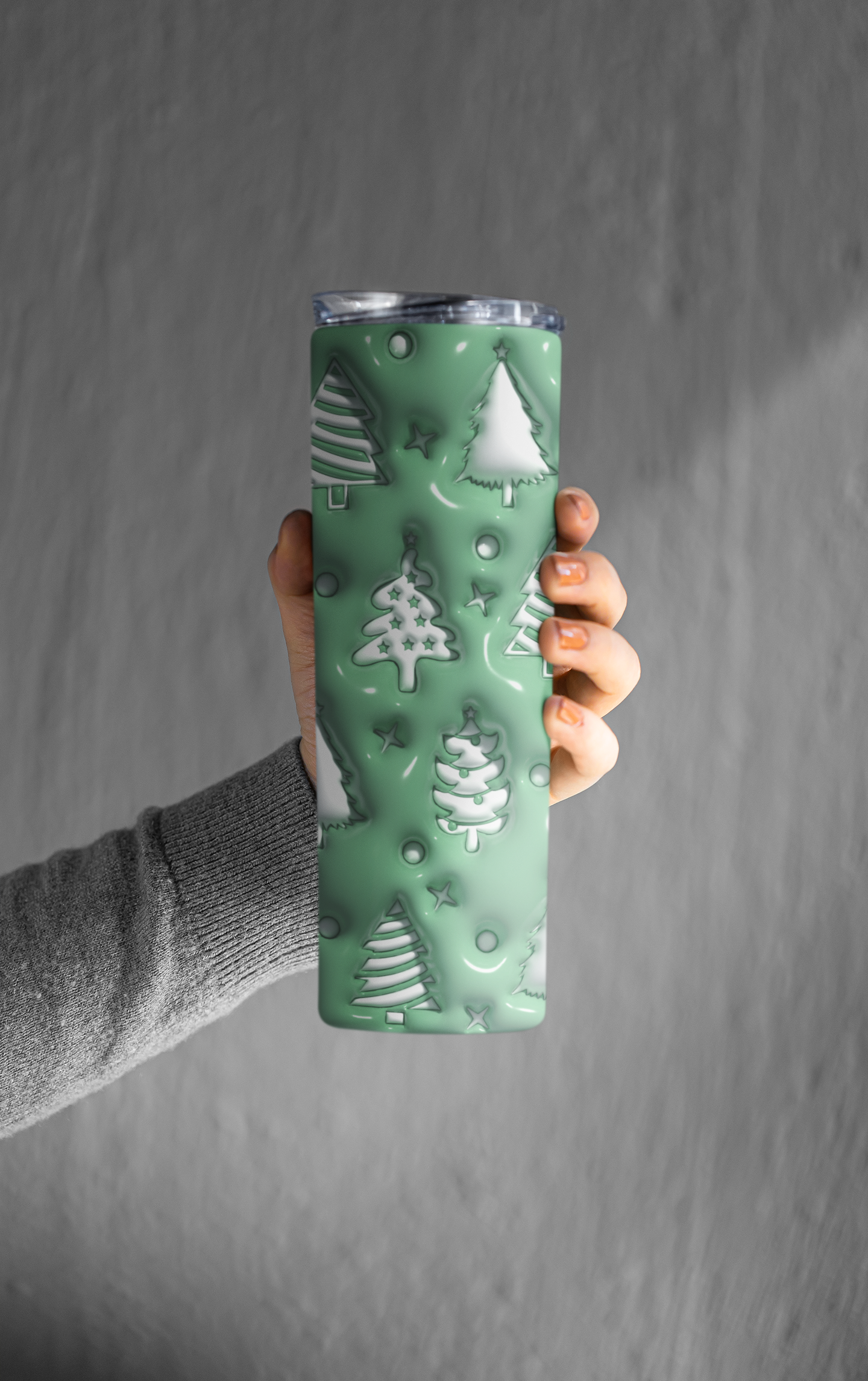 A hand holding a tall insulated tumbler with a mint green holiday design featuring embossed Christmas trees and snowflakes, set against a textured gray background.