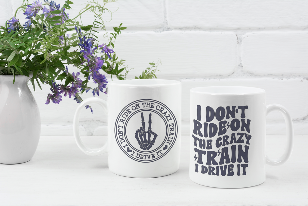 Two white ceramic mugs with dark gray printed text and a hand graphic, displayed on a white surface in front of a white brick wall with a vase of purple flowers on the left.