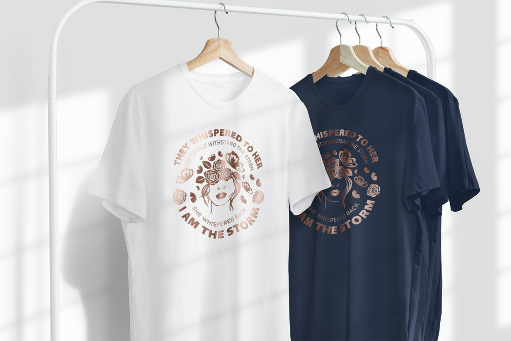 A white T-shirt and a navy blue T-shirt hanging on wooden hangers from a white clothing rack. Both shirts feature a circular graphic with a stylized dog face and the text 'THAT WHISPERED TO HER I AM THE STORM'.
