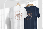 A white T-shirt and a navy blue T-shirt hanging on wooden hangers from a white clothing rack. Both shirts feature a circular graphic with a stylized dog face and the text 'THAT WHISPERED TO HER I AM THE STORM'.