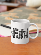 A white ceramic coffee mug with a black typographic cross design spelling 'faith' and the words 'ROMANTIC' and 'BELIEVE', placed on a wooden table with a blurred desk background.