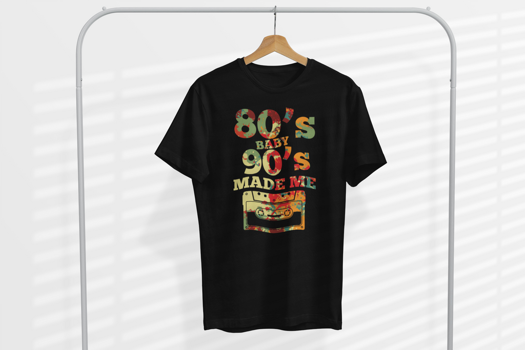 A black short-sleeve t-shirt hanging on a wooden hanger from a white clothing rack. The shirt features a colorful retro graphic on the front with the text '80's BABY 90's MADE ME' and a cassette tape illustration.