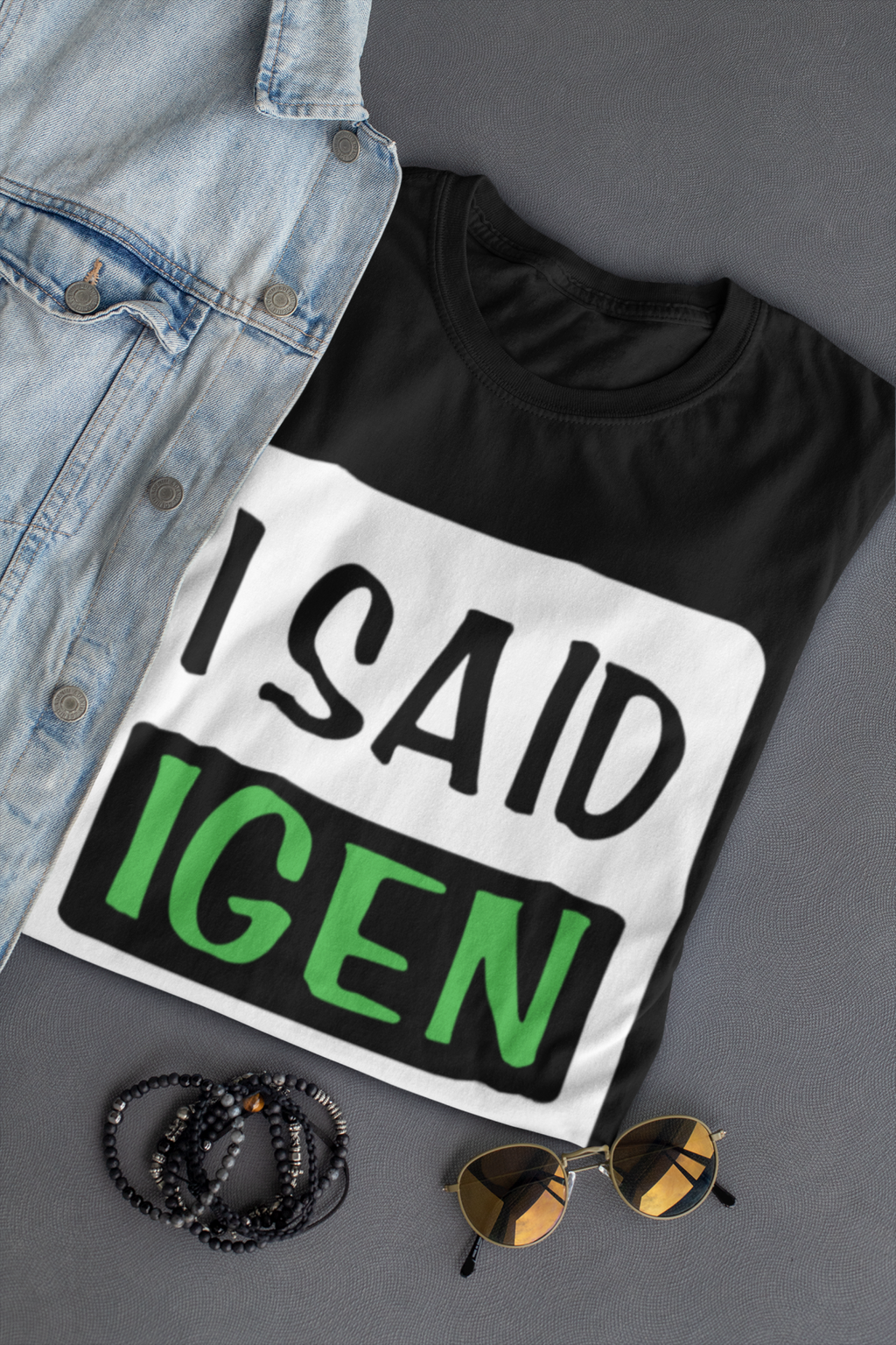 Black t-shirt with 'I SAID IGEN' text, denim jacket, sunglasses, and beads on a gray surface