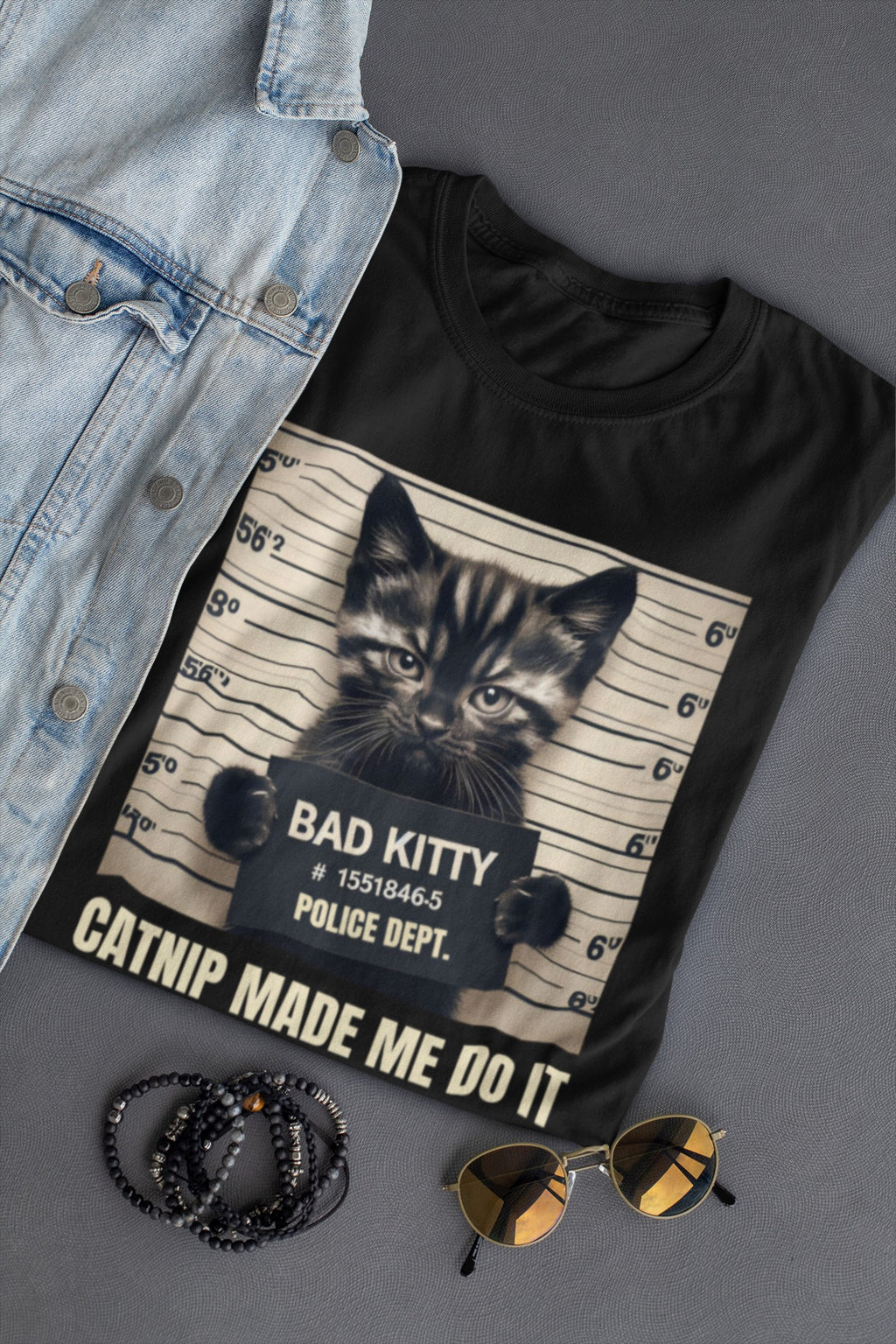 Black t-shirt with a cat graphic and text, denim jacket, sunglasses, and beads on a gray surface