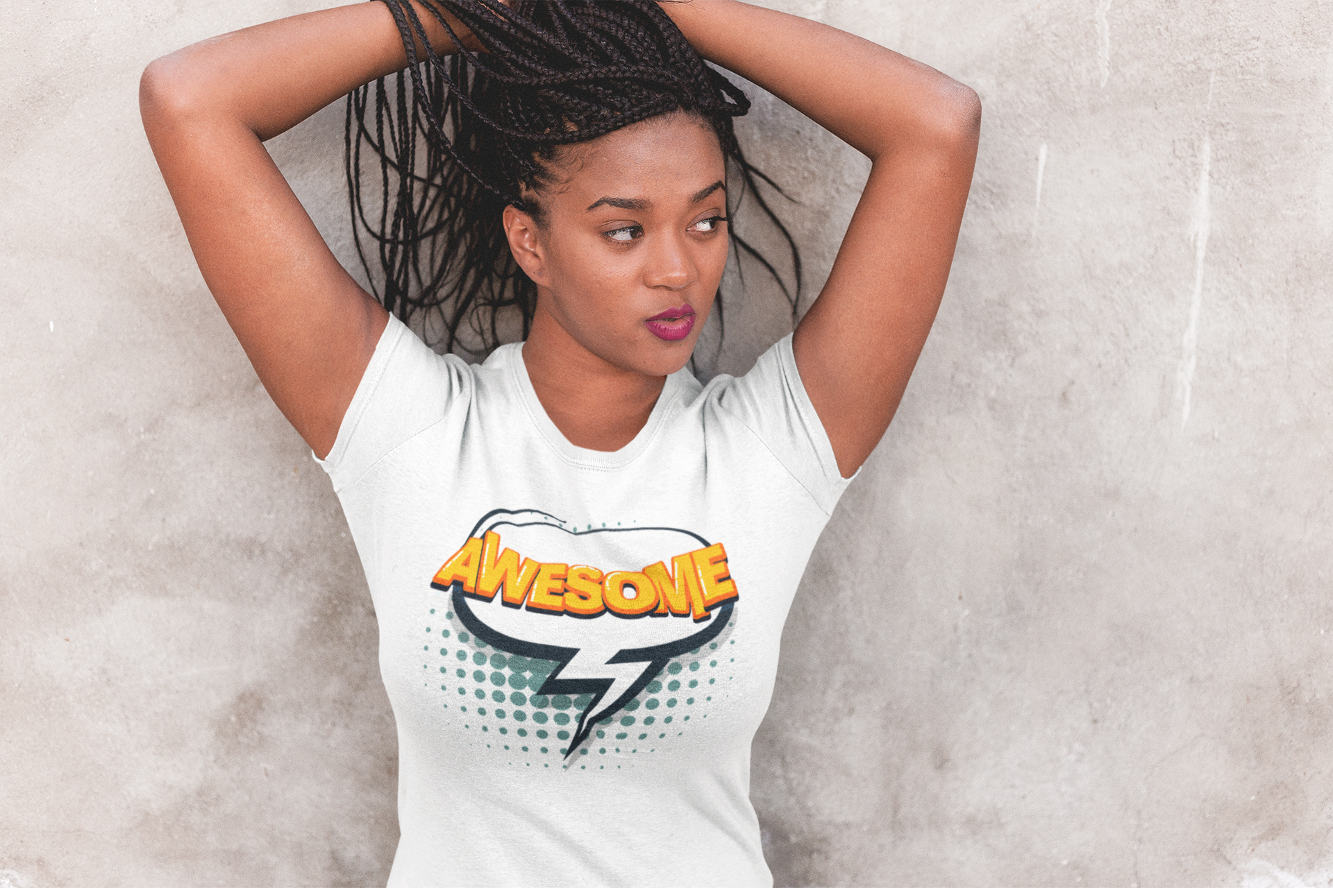 A woman with braided hair wearing a white graphic t-shirt with a comic-style 'AWESOME' print, posing against a textured gray concrete wall.