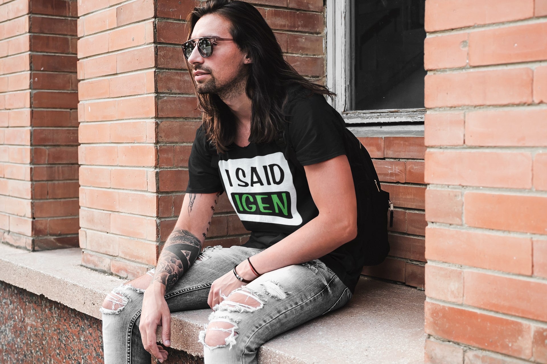 Man wearing a black t-shirt with text, sitting against a brick wall.