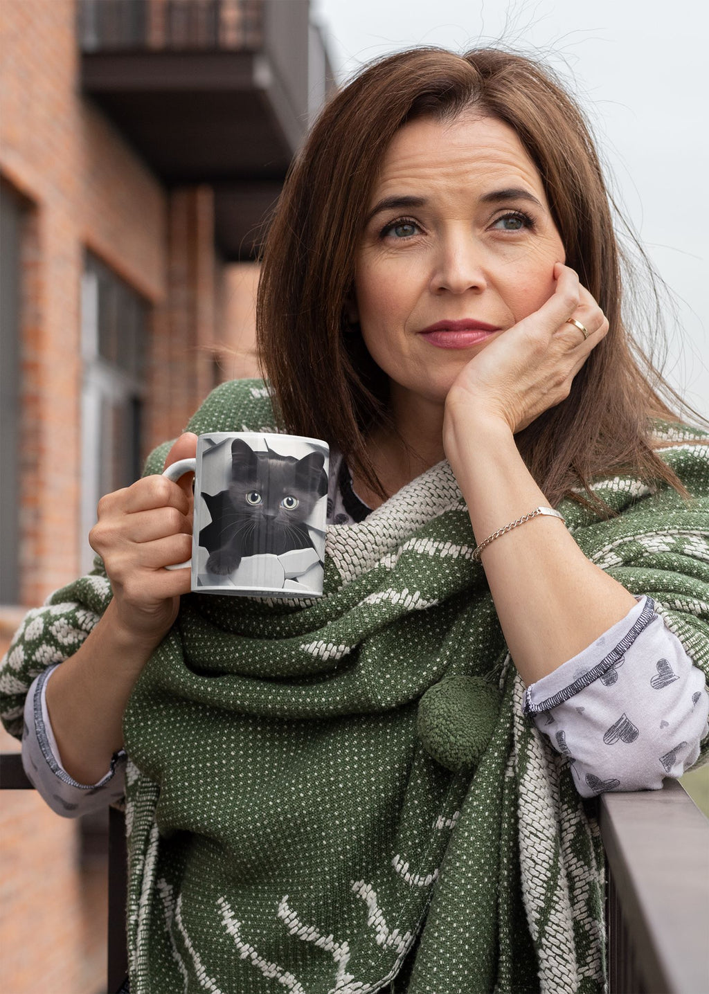 Woman holding a mug with a cat design, wearing a green patterned shawl outdoors.