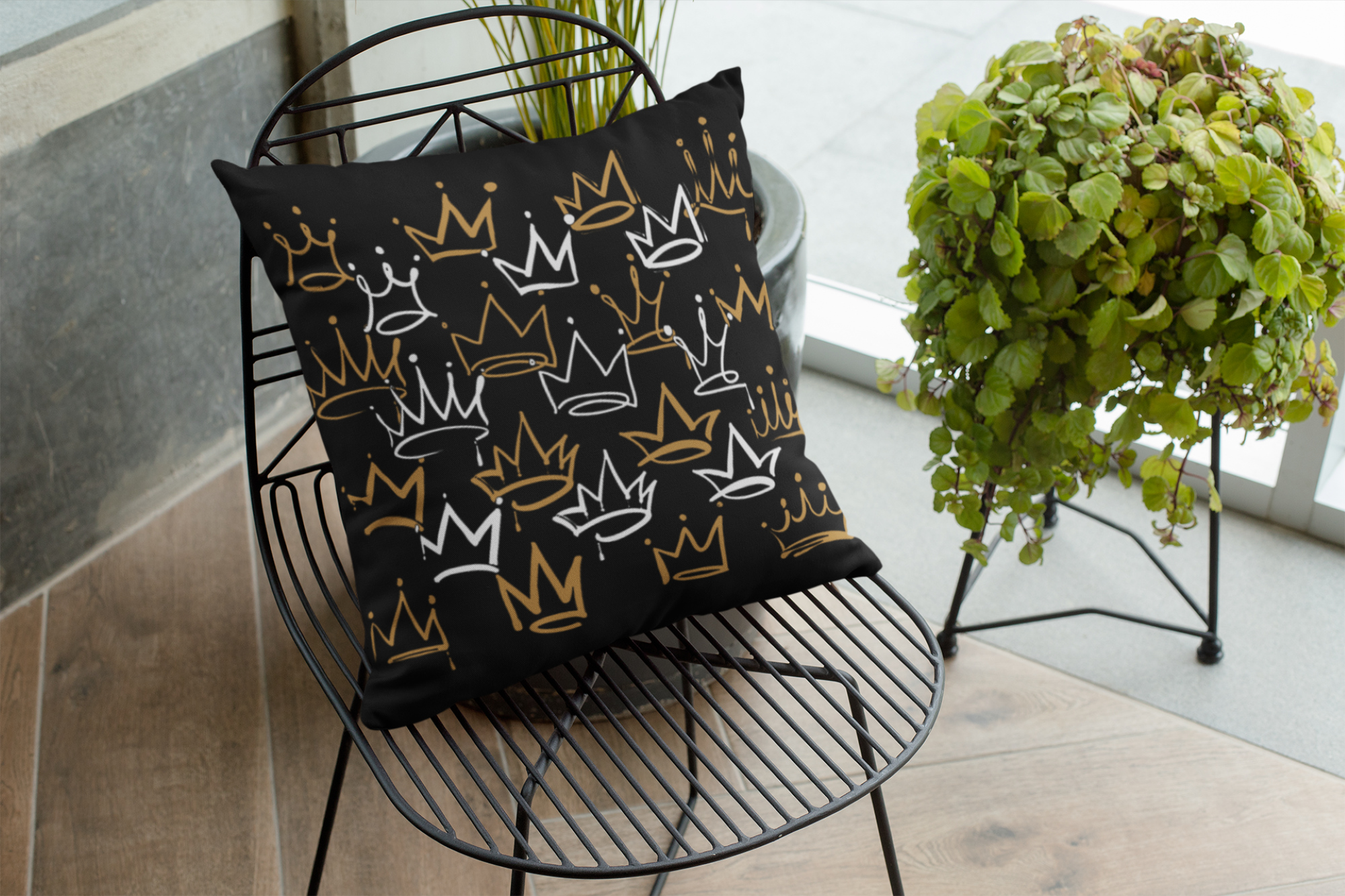 Decorative pillow with crown patterns on a black wire chair next to a potted plant.