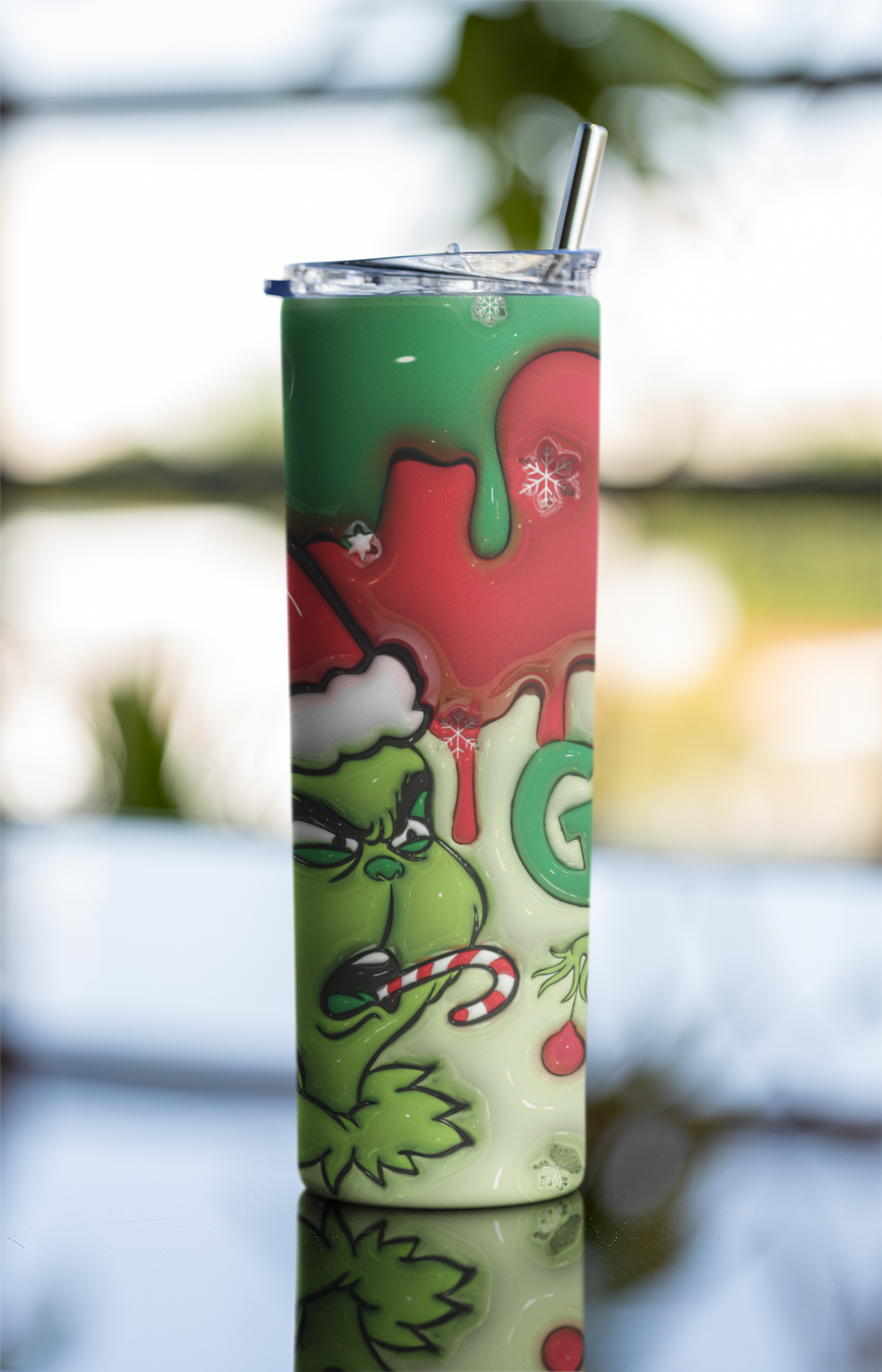 A tall, skinny tumbler with a clear lid and metal straw, featuring a Grinch-themed Christmas design with green, red, and white graphics, set against a blurred outdoor background.